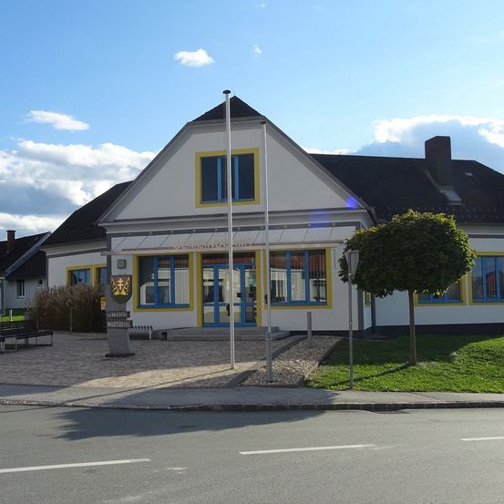 Ein weißes Gebäude mit gelber Umrandung hat mehrere Fenster und einen Fahnenmast vorn. Der Eingang befindet sich unter einem Vordach. Davor stehen Bänke und ein Baum.