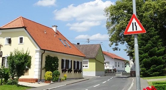 Eine kleine Stadtstraße mit einigen Häusern und einem Verkehrsschild, das eine Kreuzung anzeigt. Die Häuser haben rote Dächer und grüne Büsche davor.