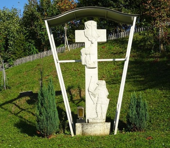 Ein weißer Kreuz auf einem Hügel, geschützt durch eine Überdachung, zeigt einen Soldaten und ein Reh. Bäume und ein Holzzaun umgeben das Kreuz.