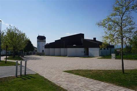 Ein leerer Parkplatz vor einem großen Gebäude mit Turm. Bäume und Gras umgeben das Gelände.