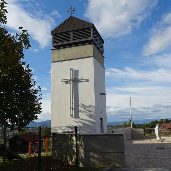 Ein hoher weißer Turm mit einem Kreuz darauf steht vor einer Kulisse von Bergen. Ein Zaun umgibt den Turm und ein kleines Gebäude steht in der Nähe.