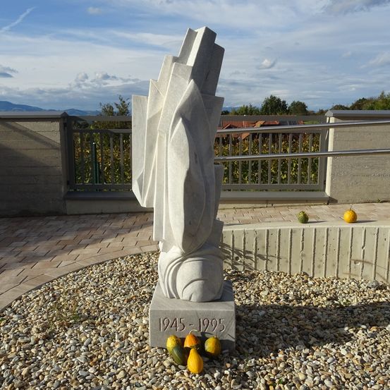 Eine abstrakte Steinskulptur mit den Jahren 1945-1995 eingraviert steht auf einem Kiesbereich. Sie ist von grünen und gelben Kürbissen umgeben, und der Hintergrund zeigt einen Zaun und eine entfernte Aussicht auf Bäume und Berge.