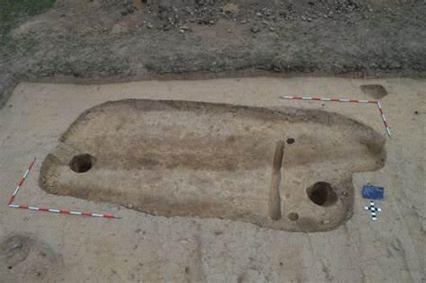 Eine Luftaufnahme einer archäologischen Ausgrabung zeigt eine freigelegte, ovalförmige Struktur mit zwei Löchern und einem kleinen blauen Objekt in der Nähe.
