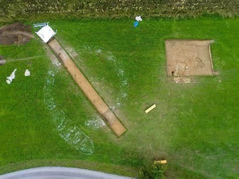 Eine Luftaufnahme einer archäologischen Ausgrabungsstätte in einem grasigen Feld mit einem Erdweg, einer Leiter und mehreren Stoffstücken, die verstreut herumliegen.