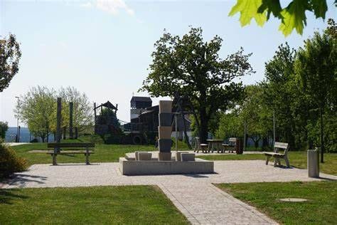Ein Freiluftpark mit Bänken, einem Springbrunnen und einem hölzernen Spielhaus, umgeben von Bäumen und Gras unter einem klaren blauen Himmel.