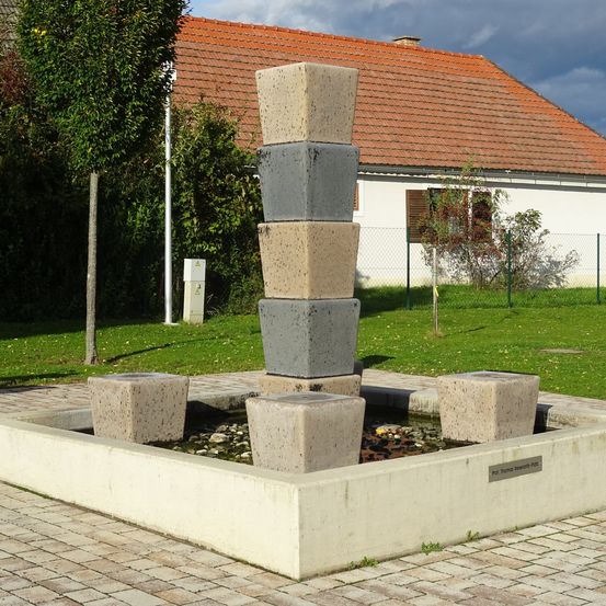 Ein Betonbrunnen mit mehreren gestapelten Blöcken und fließendem Wasser vor einem Haus mit rotem Dach.