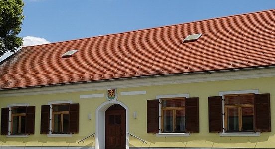 Ein gelbes Gebäude mit rotem Dach und braunen Fensterläden. Es hat eine große Tür mit einem Wappen darüber. Die Fenster sind geschlossen und ein Baum steht links.