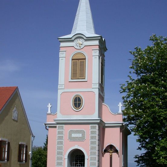 Ein rosafarbener Kirchturm mit Uhr, Kreuz und Fenster steht gegen einen blauen Himmel. Der Turm wird von einem beigefarbenen Gebäude und einem Baum flankiert.