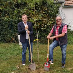 Zwei Männer stehen neben einem Baum, den sie pflanzen. Sie halten beide eine Schaufel und lächeln. Dahinter steht ein Haus mit einem Zaun.