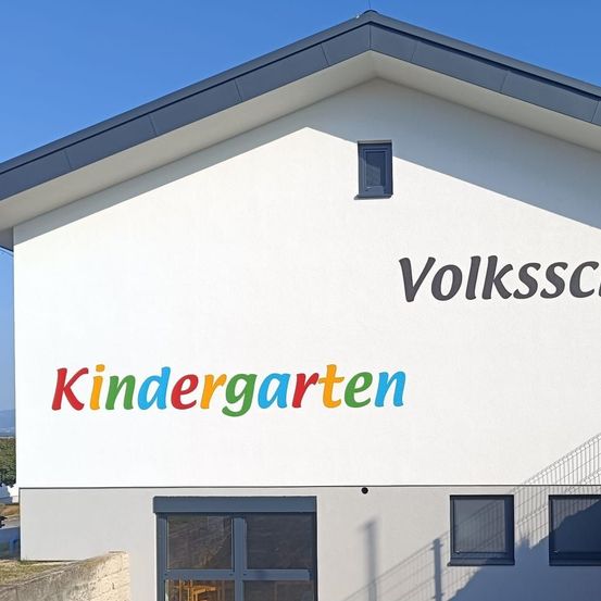Ein weißes Gebäude mit einem blauen Dach hat ein farbenfrohes Schild mit der Aufschrift 'Volksschule Kindergarten' an seiner Fassade. Das Gebäude hat drei Fenster und eine Tür.