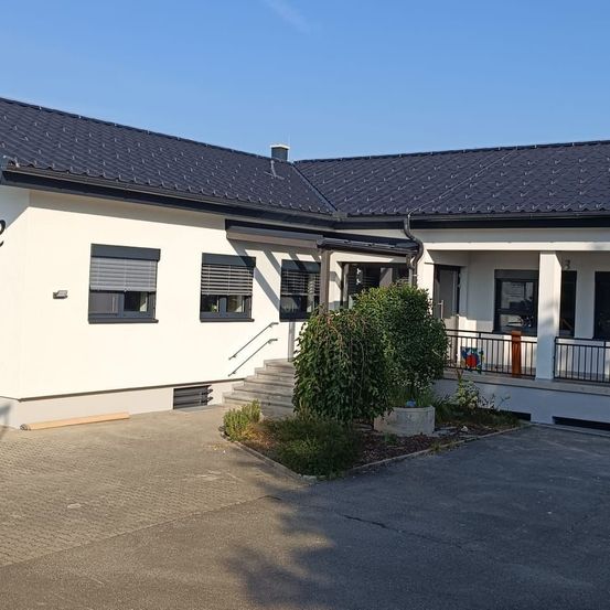 Ein modernes weißes Haus mit schwarzem Dach, ausgestattet mit drei Fenstern und einem überdachten Eingang. Es hat eine Einfahrt mit einem kleinen Garten und einem Geländer an der Seite.