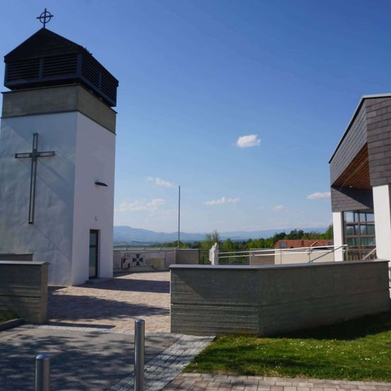 Ein weißer Kirchturm mit einem Kreuz steht vor einem blauen Himmel. Ein grasbewachsener Bereich davor mit einer Betonbarriere.