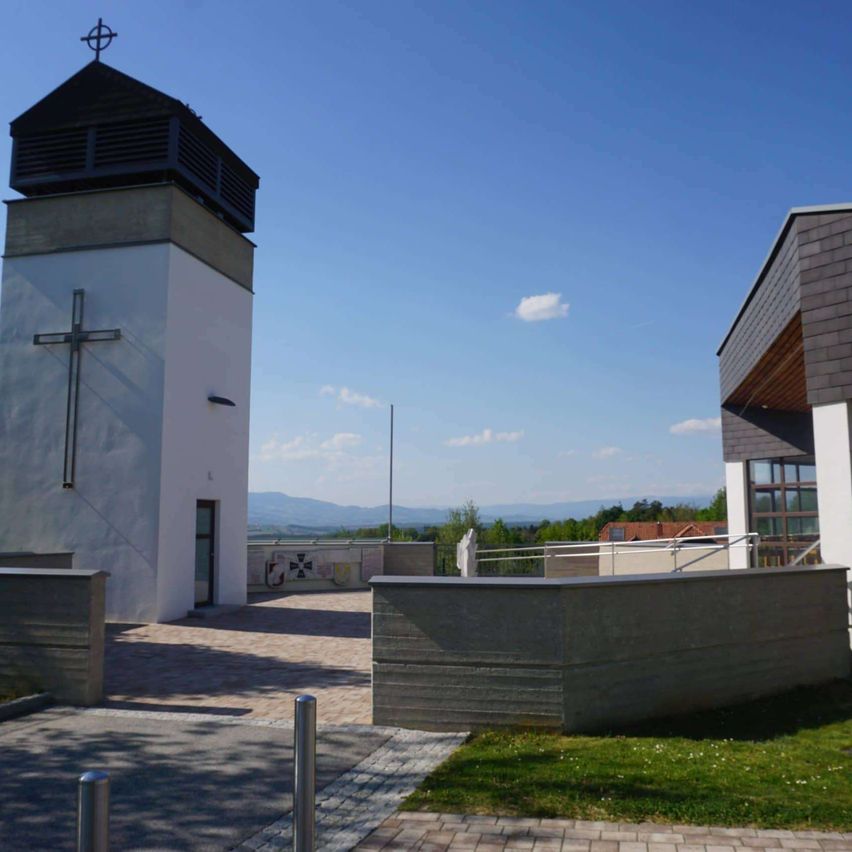 Ein weißer Kirchturm mit einem Kreuz steht vor einem blauen Himmel. Ein grasbewachsener Bereich davor mit einer Betonbarriere.