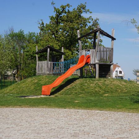 Ein Außenspielplatz mit einer roten Rutsche auf einem Hügel, umgeben von Holzspielgeräten und Grün, unter einem klaren blauen Himmel.