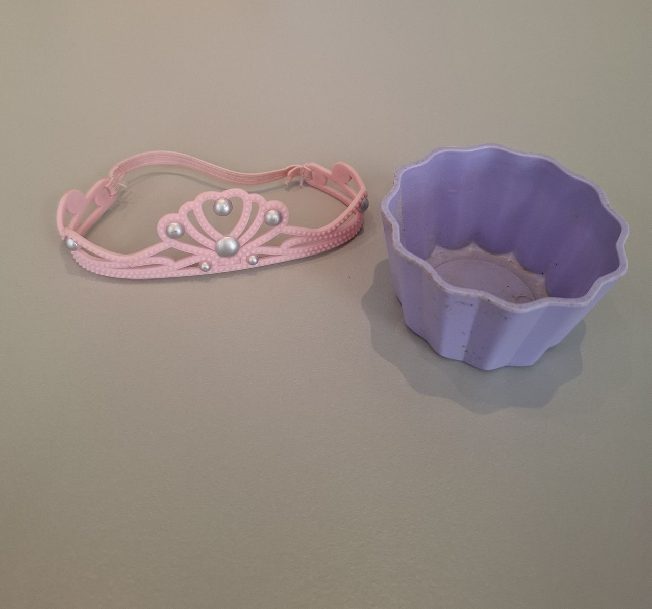 Ein rosa Kopfband mit Perlen und ein lila blumenförmiger Behälter liegen auf hellem Hintergrund.