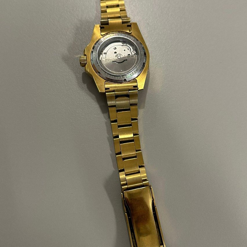 Eine goldfarbene Armbanduhr mit silbernem Zifferblatt wird vor einem grauen Hintergrund gezeigt. Die Uhr hat ein mechanisches Uhrwerk, das durch den transparenten Gehäuseboden sichtbar ist.