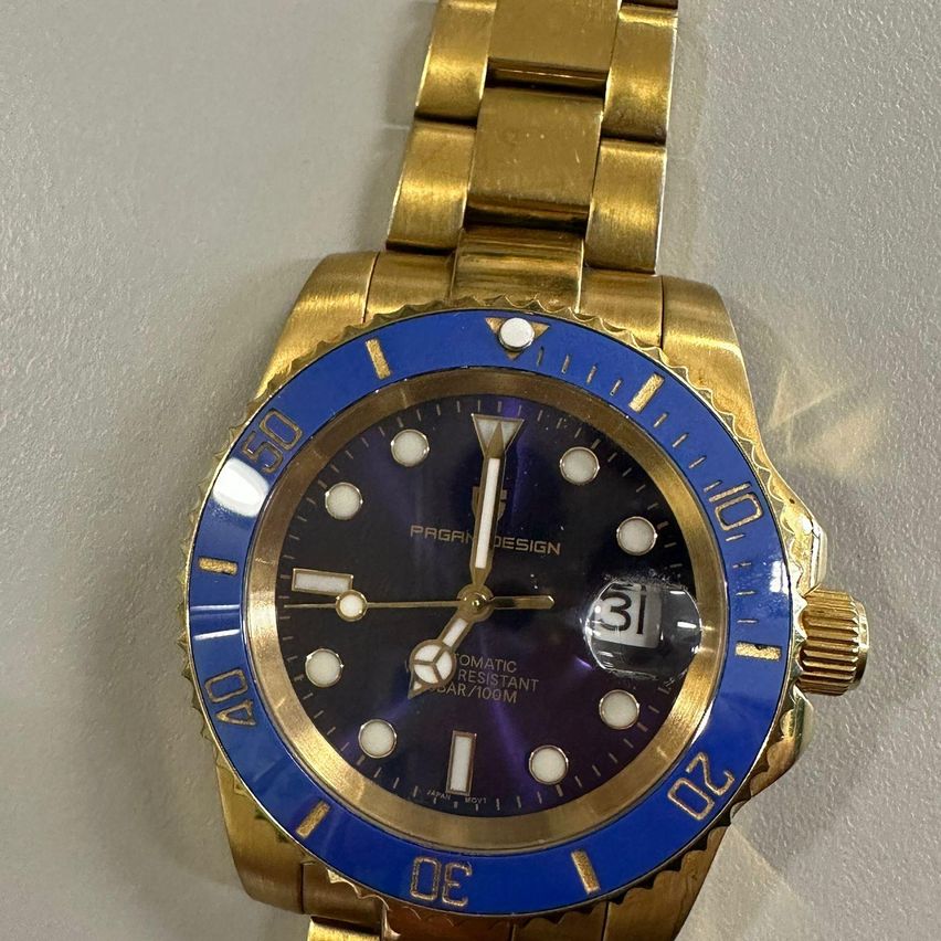 Eine goldene Armbanduhr mit blauem Zifferblatt, weißen Zeigern und Markierungen sowie schwarzem Lünettenring mit goldenen Akzenten. Die Uhr zeigt 'PAGRIN DESIGN' und 'OMATIC RESISTANT GAR7106M' auf dem Zifferblatt.