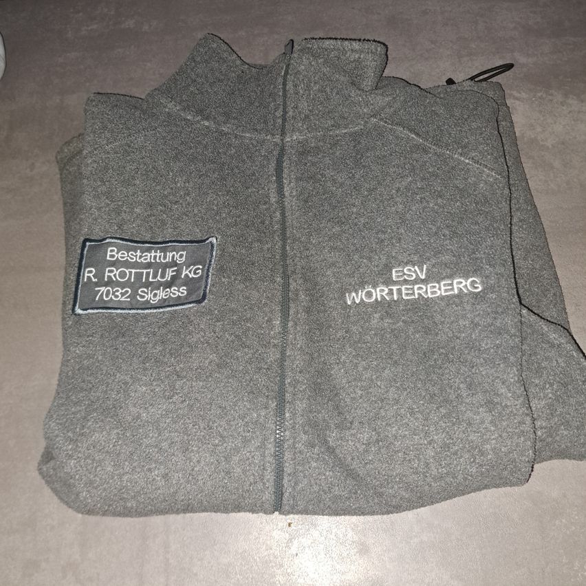 A gray fleece jacket with a zipper. It has a label with 'Bestattung R. Rottluff KG' and another label with 'ESV Worterberg'. The jacket is folded and placed on a surface.