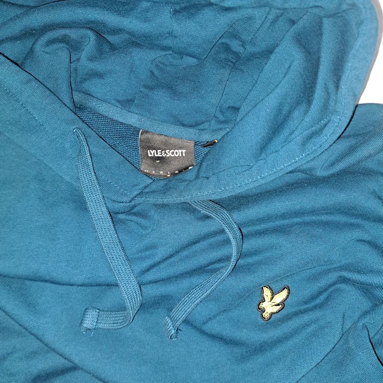 A close-up of a teal hoodie with a Lyle & Scott label on the left side and a yellow bird patch on the right side.