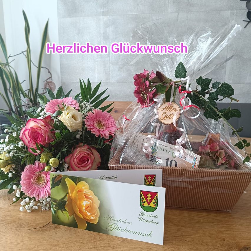 A bouquet of pink and white flowers and a gift basket with a wine bottle and other items on a wooden table. The basket has a tag with the words 'Alles Gute'. The bouquet has a yellow rose and other flowers. A card is also on the table.