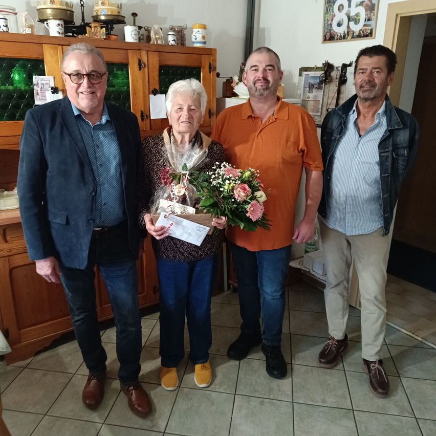 Four adults stand in a room. An elderly woman holds a bouquet of flowers and a gift box. The man on her left wears a suit. The man in the middle wears an orange shirt. The man on the right wears a denim jacket.