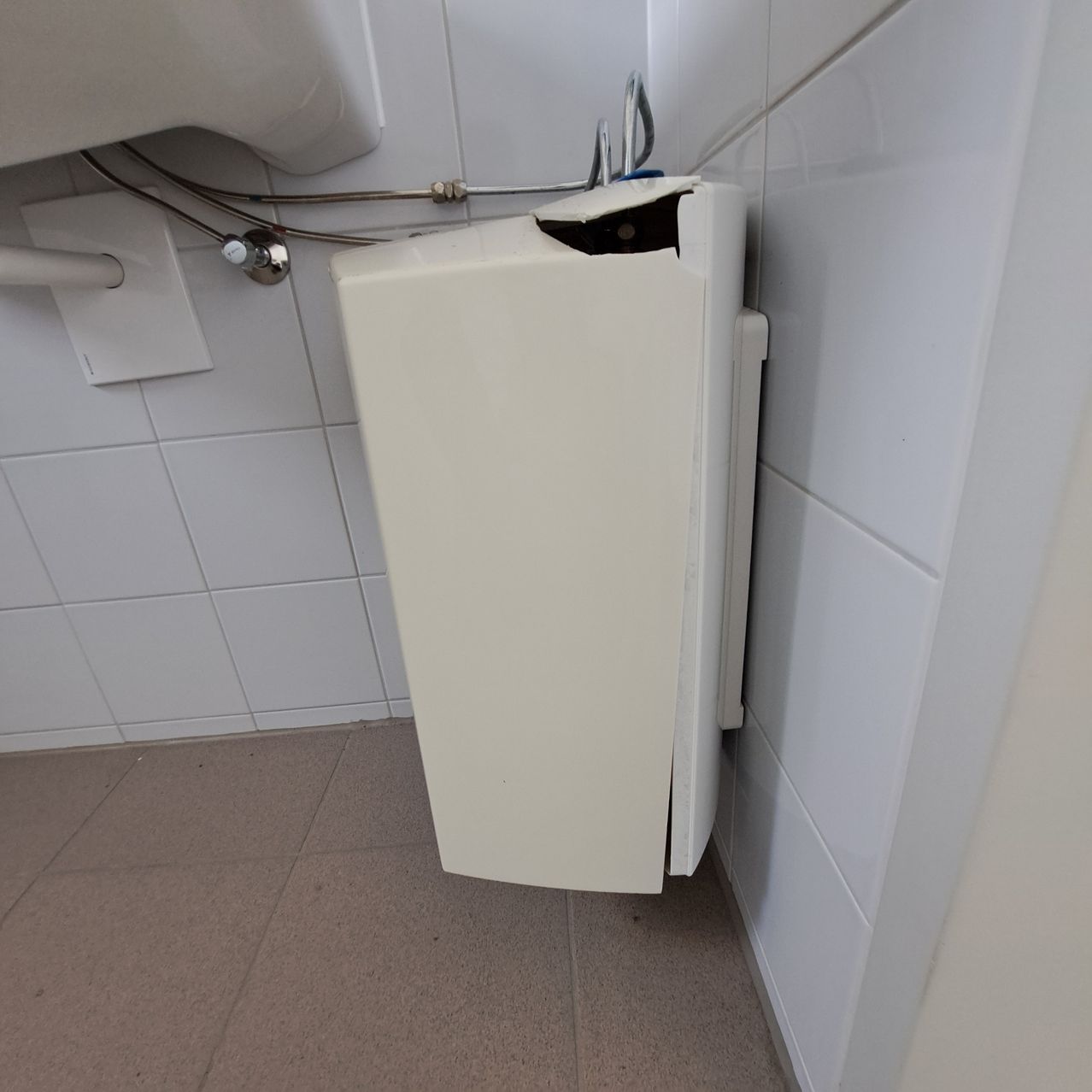 A white soap dispenser is mounted on a tiled wall in a bathroom, with a broken top and a silver faucet nearby.