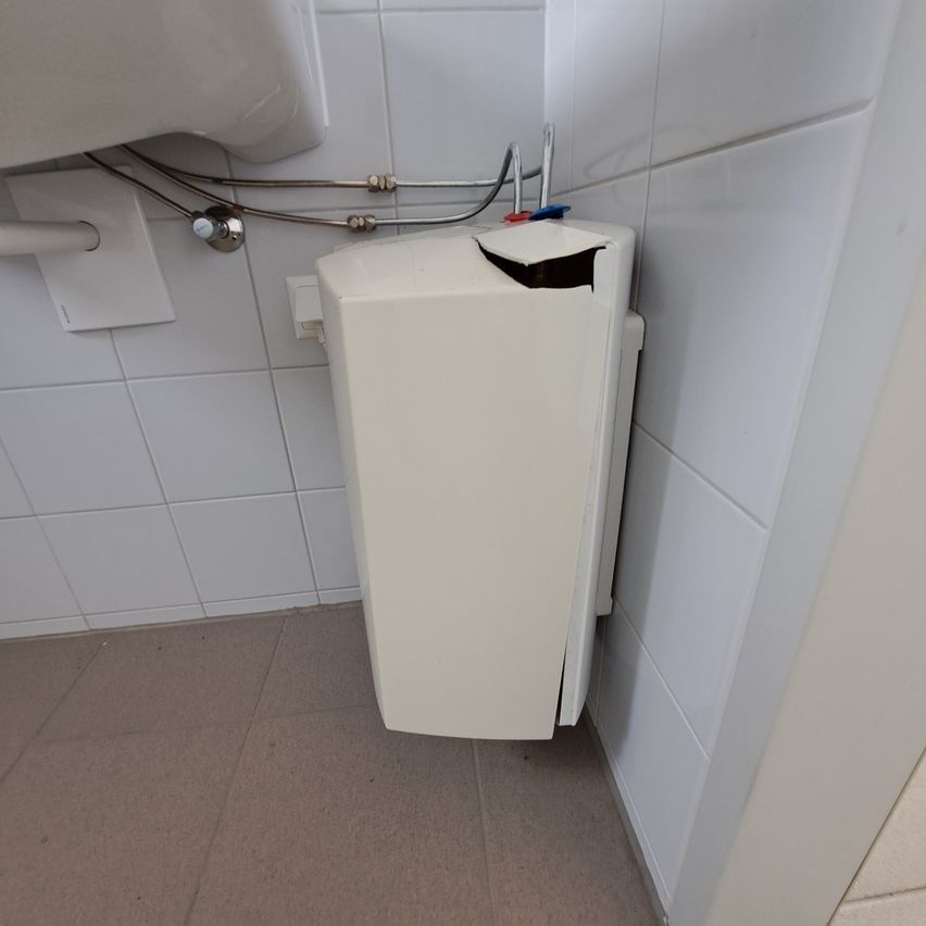 A white toilet paper dispenser with a broken cover is mounted on a tiled wall in a bathroom.