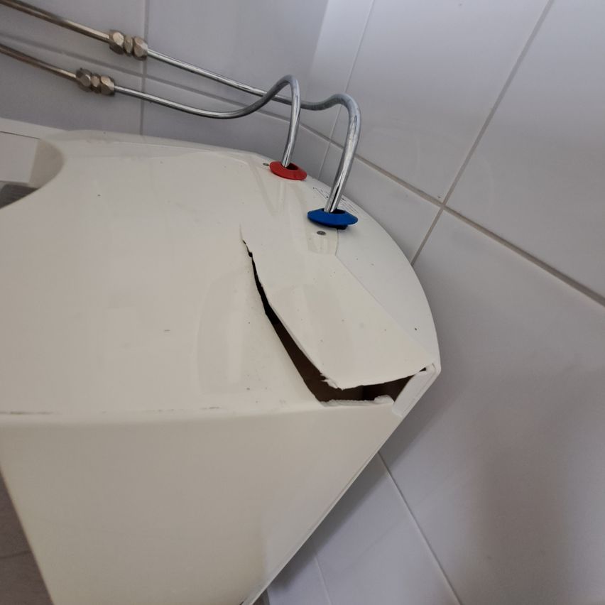 A damaged white water heater with metal pipes attached, mounted on a tiled wall.