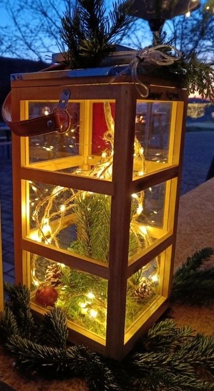 A wooden lantern with a Christmas tree and lights inside. It is on a wooden table with pine branches and an apple. The lantern has a leather strap and is placed in front of a blurry background.