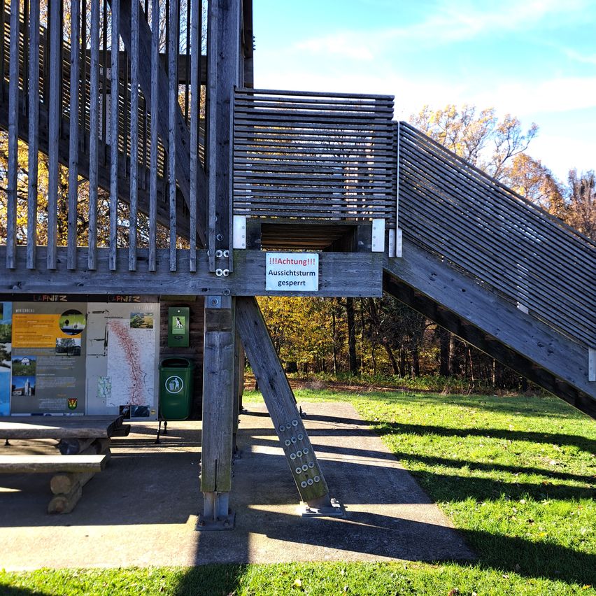 A wooden staircase with a handrail and a sign warning of storm danger. Adjacent to the stairs is a wooden bench and a trash can. There is a board with information. In the background, trees with yellow leaves.