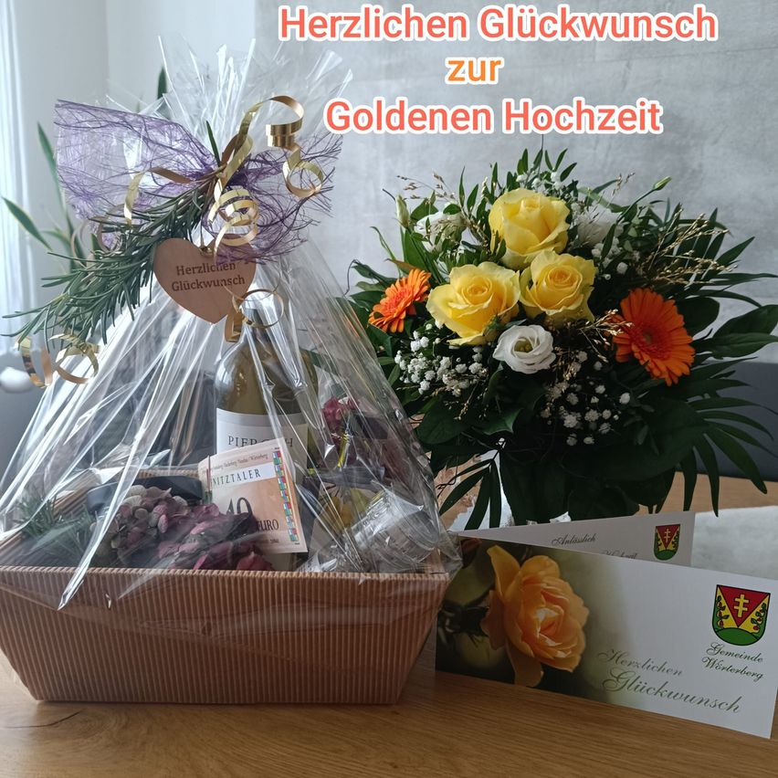 Ein Geschenkkorb mit einer Weinflasche und einem Strauß gelber und orangefarbener Blumen mit einem Schild, das 'Herzlichen Gluckwunsch zur Goldenen Hochzeit' auf einem Holztisch steht.