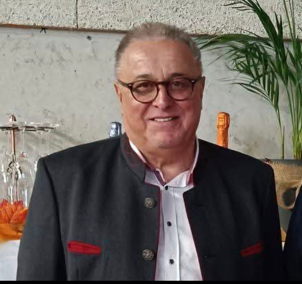 Ein lächelnder Mann mit Brille und einer schwarzen Jacke mit roten Akzenten steht vor einer weißen Wand mit einer Pflanze und Weingläsern.