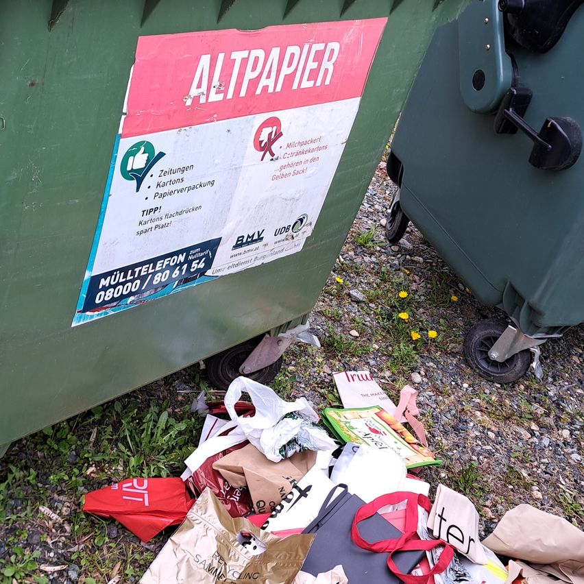 A green garbage bin with a sign that reads "Altpapier". The sign includes a phone number and a green recycling logo. The ground is littered with various items.