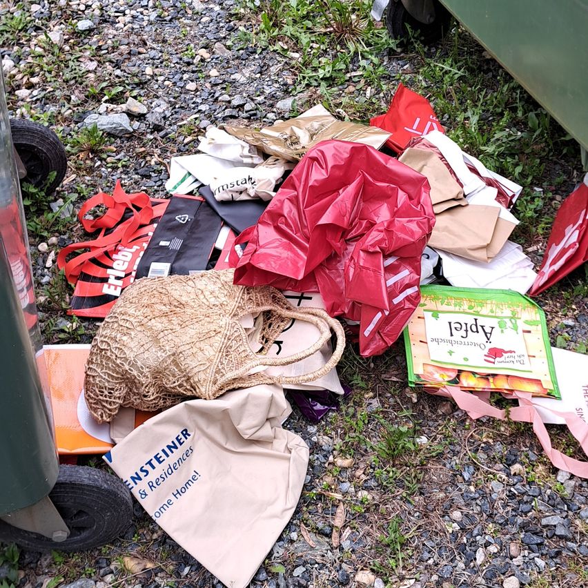 A pile of discarded bags and trash is on the ground near a green dumpster, including a red bag, a box with peaches, and a brown bag.