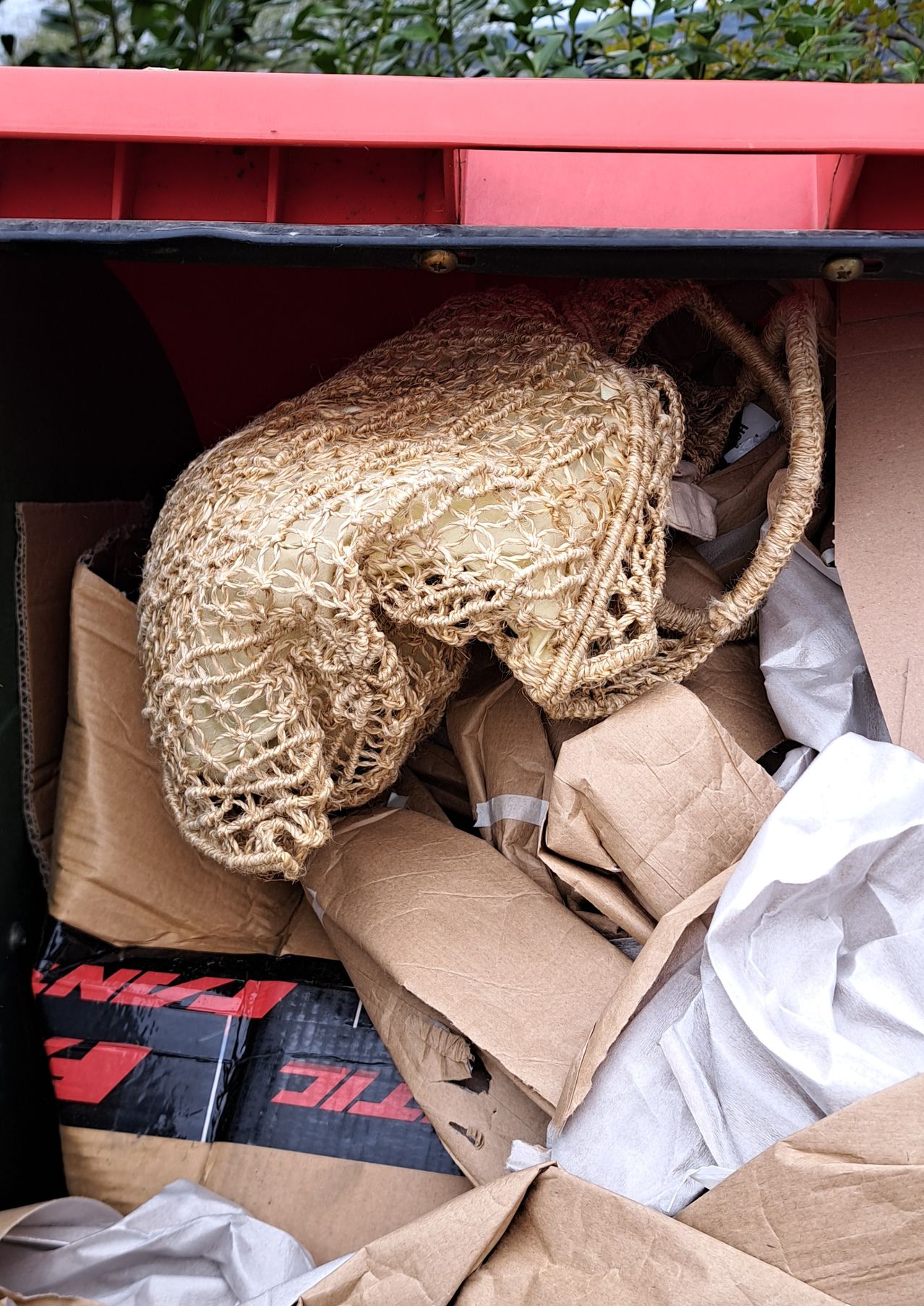A beige woven bag with handles lies amidst a pile of brown cardboard boxes and white plastic bags, possibly in a dumpster.