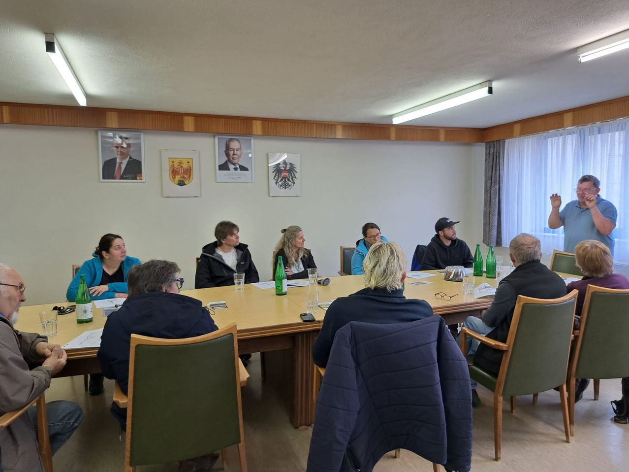 A meeting in a conference room with eight people sitting around a table. There are papers, glasses, and bottles on the table. Portraits of individuals and flags are on the wall.