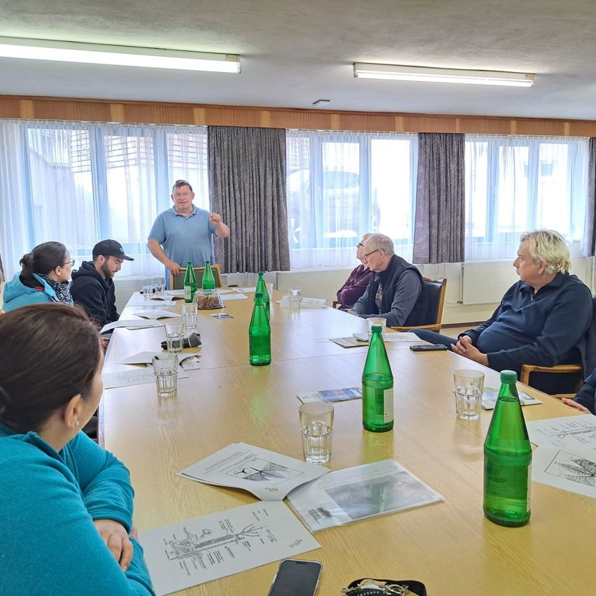 A group of people are gathered around a long table in a room with windows. A man stands in front of them, likely leading a meeting. Papers and green bottles are on the table.