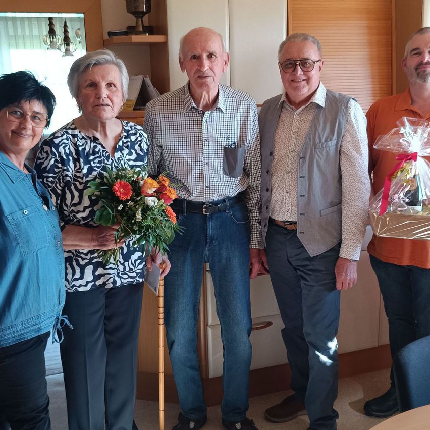 Fünf Menschen stehen in einem Raum zusammen. Ein älteres Paar hält Blumen. Ein älterer Mann hält einen Geschenkkorb. Ein weiterer Mann mit Brille steht neben ihnen. Dahinter steht ein Holzschrank.