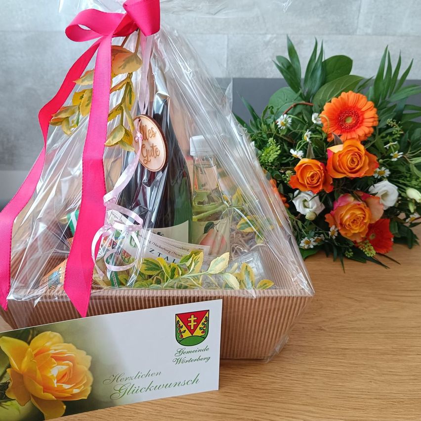 Ein Geschenkkorb mit einer Flasche Wein und Blumen, in Plastik mit einer rosa Schleife verpackt, auf einem Holztisch mit einem Strauß orangefarbener und weißer Blumen.