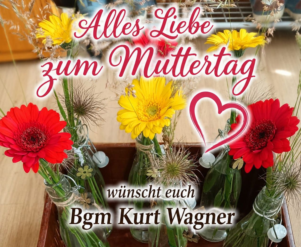 Eine Box mit Vasen mit Blumen, einschließlich roter und gelber Gerbera-Gänseblümchen, arrangiert für eine Muttertagsbotschaft von Bgm. Kurt Wagner.