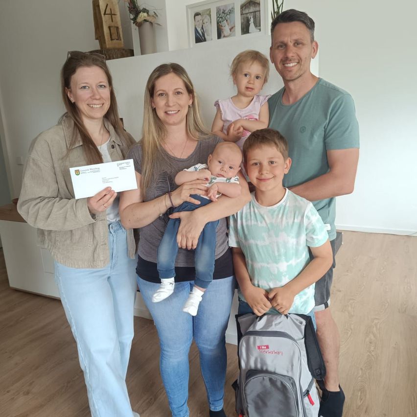 Eine vierköpfige Familie posiert für ein Foto. Eine Frau hält ein Baby, eine andere einen Scheck, und zwei Kinder stehen neben ihnen, eines mit einem Rucksack.