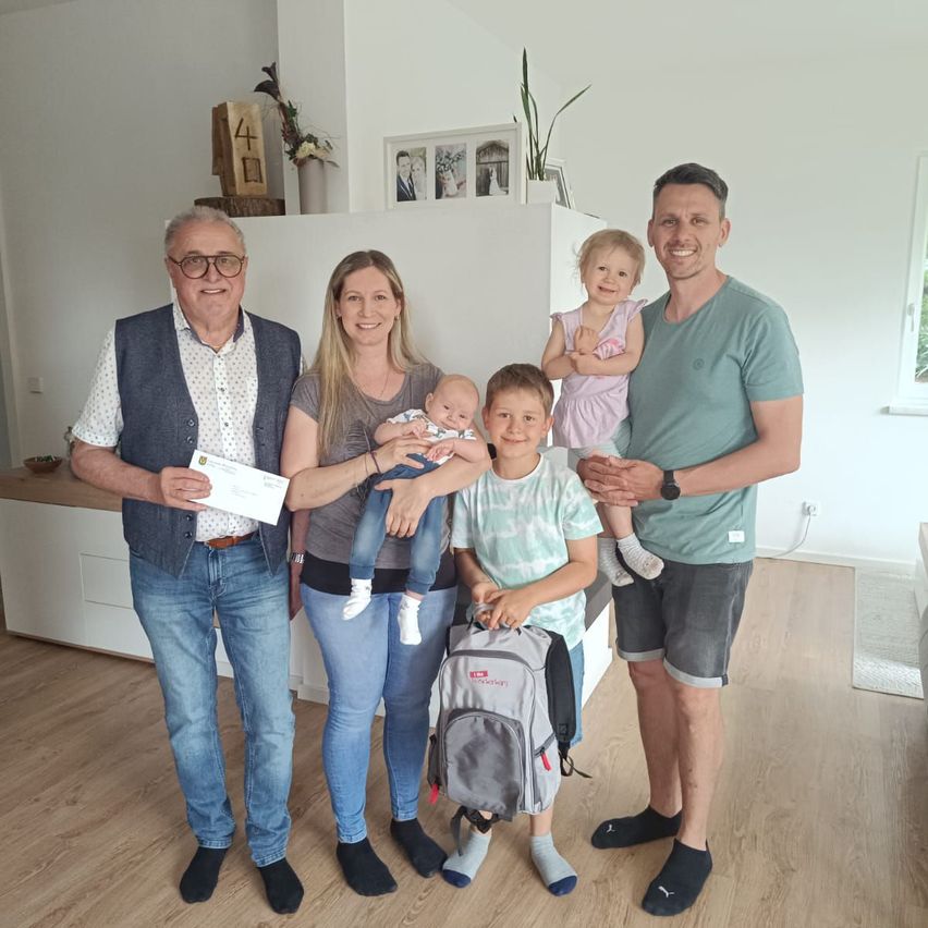 Eine Familie mit fünf Mitgliedern, darunter zwei Erwachsene und drei Kinder, steht im Innenbereich. Der Mann hält ein Papier, und die Frau trägt ein Baby. Die Kinder, eines mit Rucksack, scheinen glücklich zu sein.