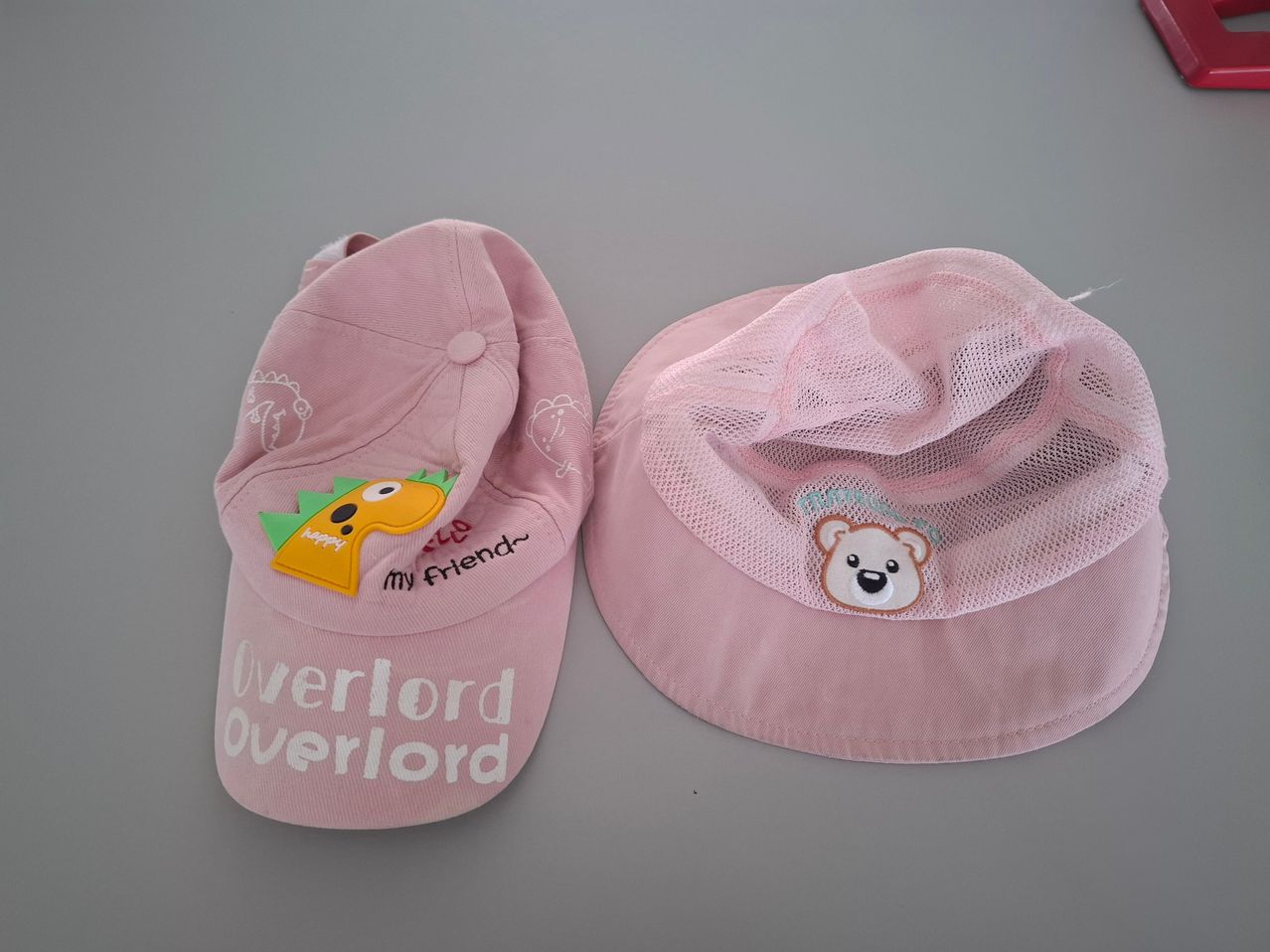 Zwei rosa Mützen sind zu sehen, eine mit 'Overlord Overlord' Text und einem Dinosaurier, die andere mit einem Bären und Netzstoff.