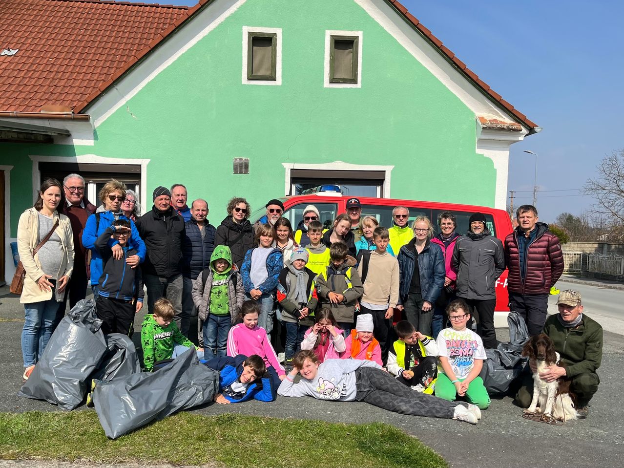 Eine Gruppe von Menschen posiert für ein Foto vor einem grünen Haus. Einige tragen Jacken, Brillen und Mützen. Es gibt auch Müllsäcke und einen Hund.