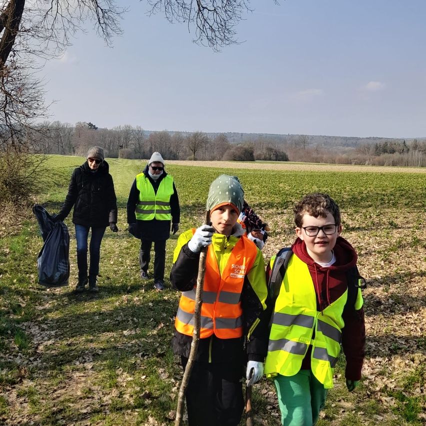 Eine Gruppe von Personen, darunter zwei Kinder in Reflektorwesten, geht auf einem Grasfeld. Sie halten einen Stock und einen Müllsack. In der Ferne befinden sich Bäume und ein weites Feld.
