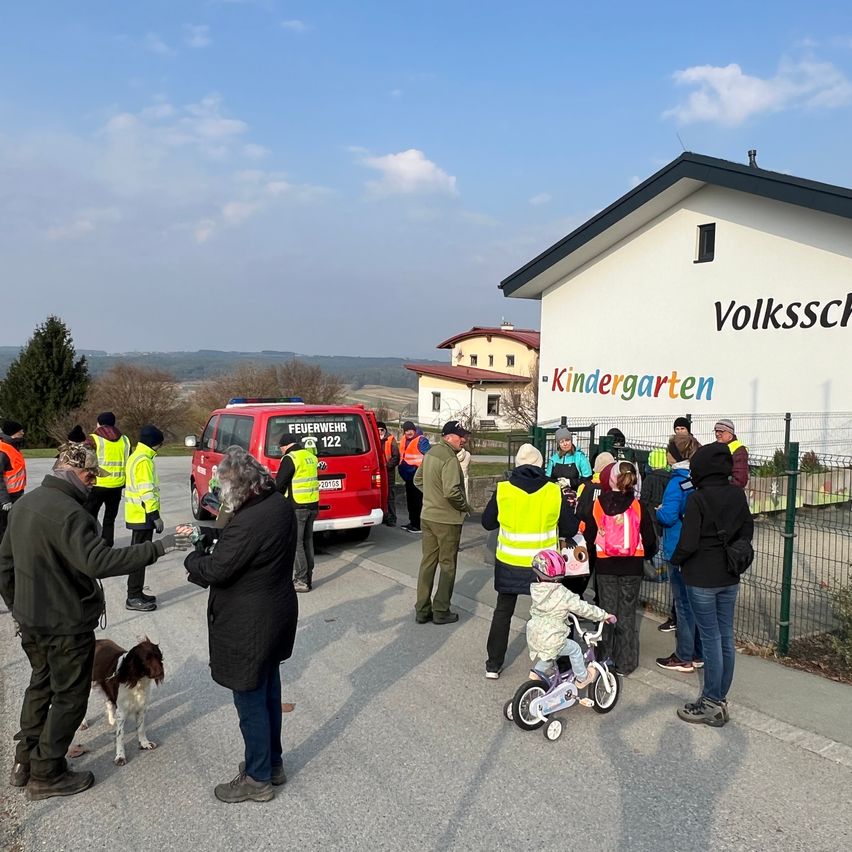 Eine Gruppe von Menschen in Sicherheitswesten steht vor einem Gebäude mit dem Wort Kindergarten. Ein Kind fährt ein Fahrrad. Ein Hund geht neben einem Mann. Ein roter Van ist geparkt.