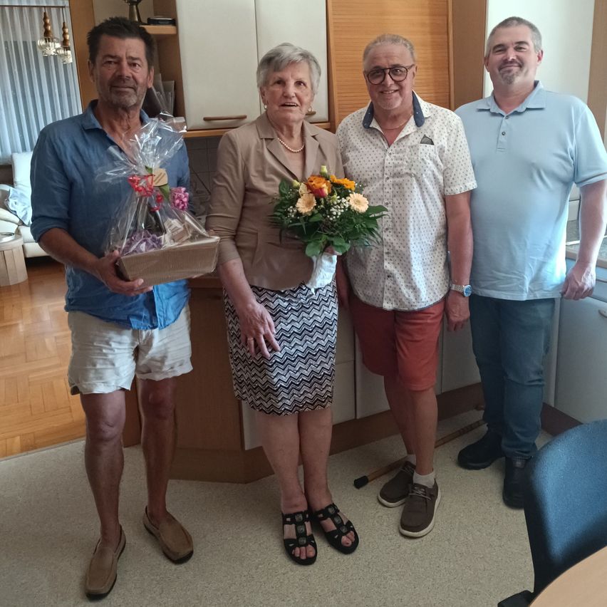 Vier Menschen stehen lächelnd in einem Raum. Eine Frau hält einen Blumenstrauß. Der Mann auf der linken Seite hält eine Geschenkbox. Dahinter befindet sich eine Küche mit Schränken und einem Tisch.
