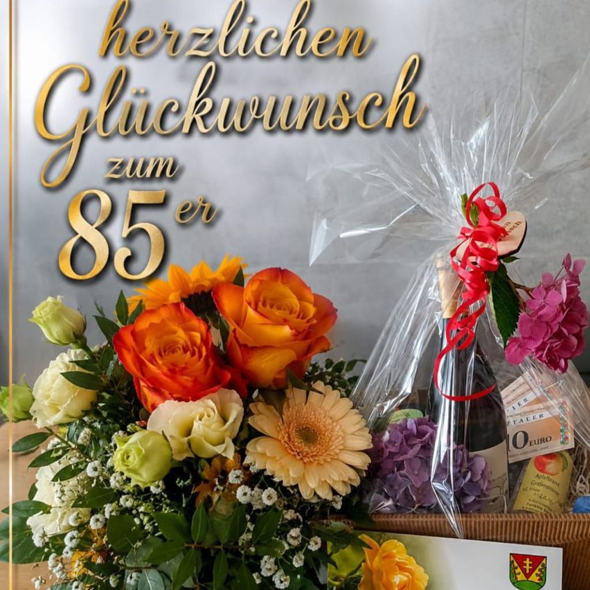Ein Blumenstrauß und eine Flasche Wein in einem Korb mit einer Schleife und einer Karte, mit den Worten 'Herzlichen Glückwunsch zum 85er' auf einem grauen Hintergrund.
