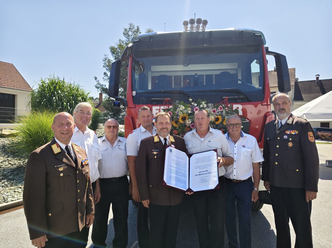 Eine Gruppe von Männern in Uniformen, einige halten ein Zertifikat, stehen vor einem roten Feuerwehrauto.