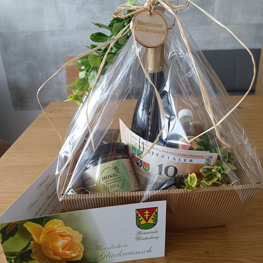 Ein Geschenkkorb mit einer Flasche Wein, einem Glas Honig und einer Karte mit einer Rose, in Plastik verpackt und mit einem Band gebunden.
